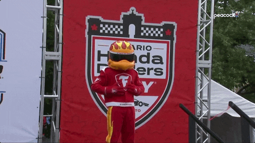 a rio honda flyers mascot stands in front of a peacock tv sign