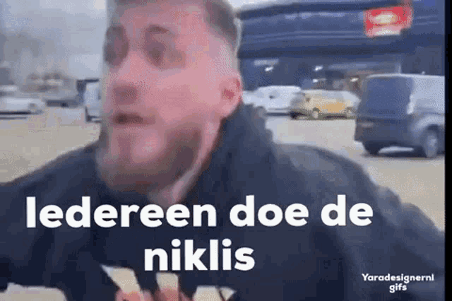 a man with a beard is standing in a parking lot with a car in the background and says iedereen doe de niklis