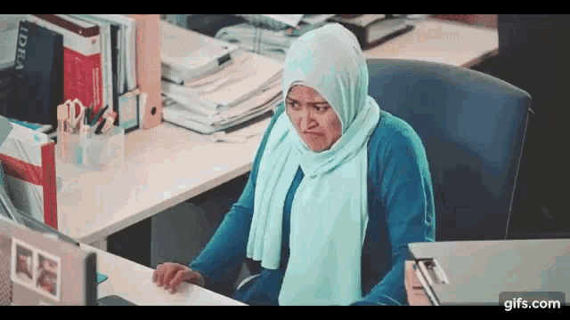 a woman wearing a hijab sits at a desk in front of a laptop