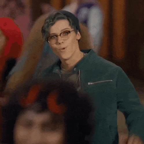 a young man wearing glasses and a green jacket is waving his hand in a crowd of people .
