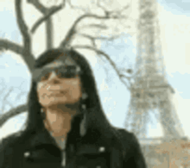 a woman wearing sunglasses and a black jacket is standing in front of the eiffel tower .