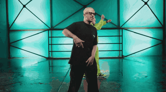 a man in a black t-shirt is dancing in front of a green wall