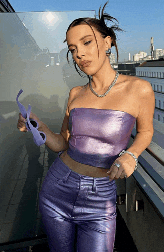 a woman wearing a purple top and purple pants holds a pair of sunglasses