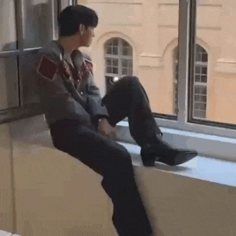 a man is sitting on a window sill with his legs crossed and looking out the window .