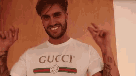 a man with a beard is wearing a gucci t-shirt and making a heart shape with his hands .