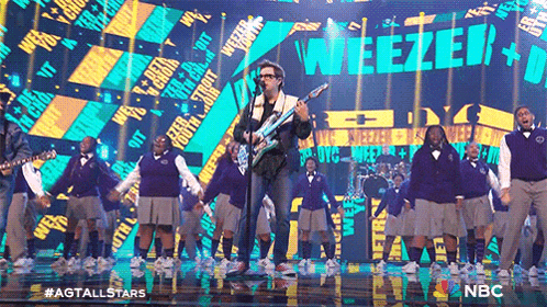 a man singing into a microphone in front of a crowd with weezer on the screen
