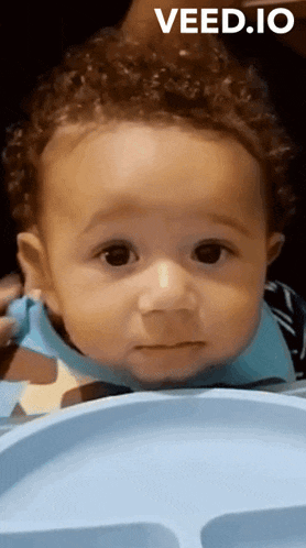 a baby with curly hair is sitting on a blue tray with the words veed.io above it .