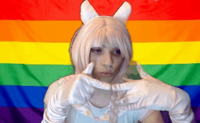 a woman in a white wig and white gloves stands in front of a colorful rainbow flag