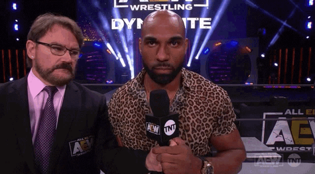 a man in a leopard print shirt holds a microphone in front of a sign that says all elite wrestling