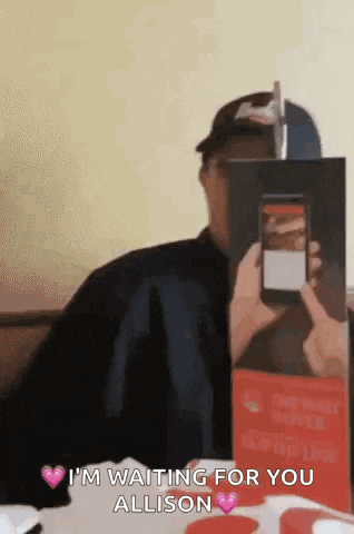 a man is holding a cell phone in front of his face and says i 'm waiting for you allison on it .
