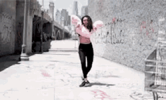 a woman in a pink top and black pants is dancing in front of a wall with graffiti on it