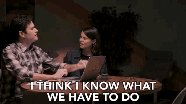 a man and a woman sit at a table with a laptop and the words " i think i know what we have to do