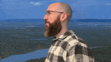 a man with a beard wearing glasses stands in front of a river