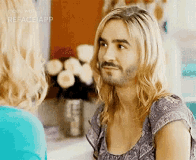a man with long blonde hair and a beard is talking to a woman in front of a vase of flowers .