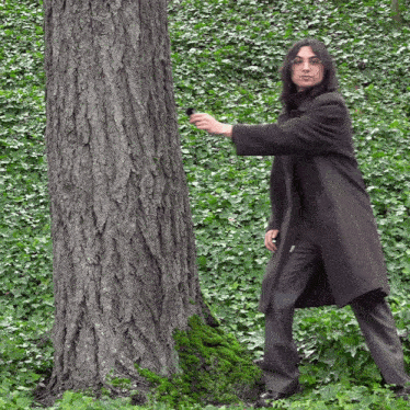 a man in a trench coat is standing next to a tree in the woods