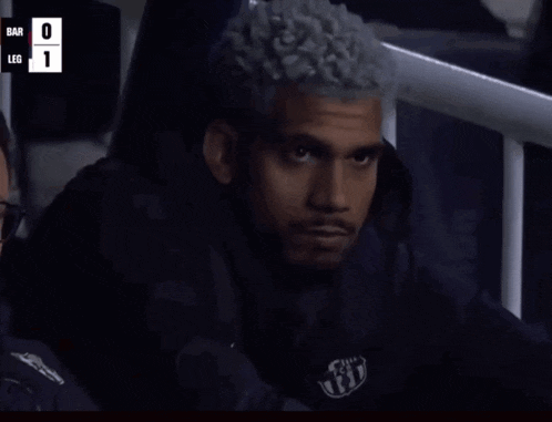 a man with curly hair is sitting in a stadium watching a game with the score bar 0 leg 1