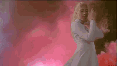 a woman in a white dress is dancing in front of a pink background with smoke coming out of her hair .