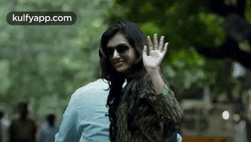 a woman wearing sunglasses is waving at a man while walking down a street .