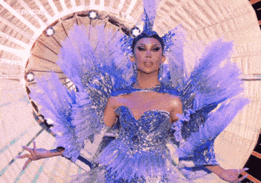 a drag queen is wearing a blue and silver dress with feathers and a crown .