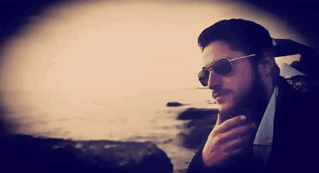 a man with a beard wearing sunglasses is standing on a rock near the ocean .