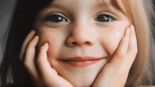 a little girl is smiling with her hands on her face and looking at the camera .