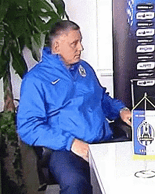 a man in a blue jacket is sitting at a table in front of a sign that says ' budimir '