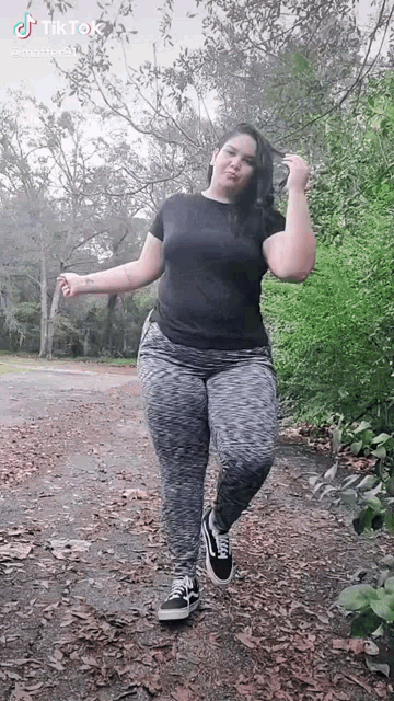 a woman in a black shirt and gray pants is standing on a dirt road in a park .
