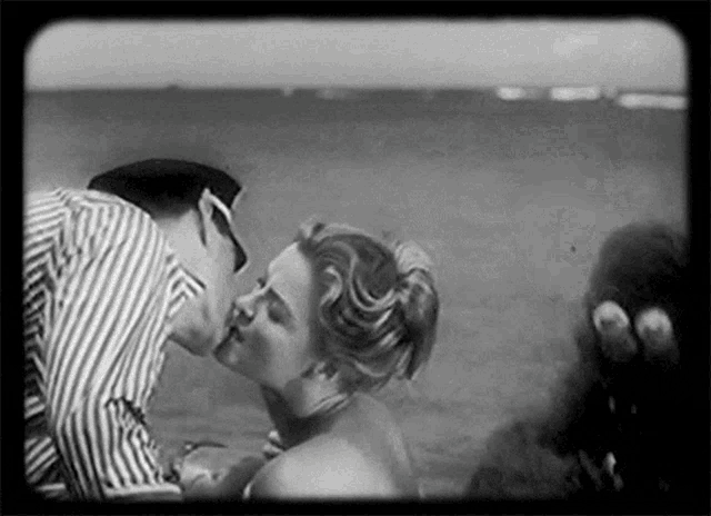 a black and white photo of a man and woman kissing on a beach