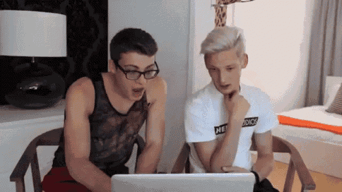 two young men are looking at a laptop screen and one has a shirt that says he