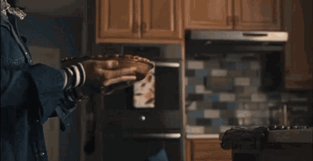 a man in a denim jacket is standing in a kitchen opening the oven door .