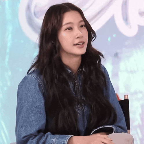 a woman with long hair is wearing a denim jacket and sitting in a chair