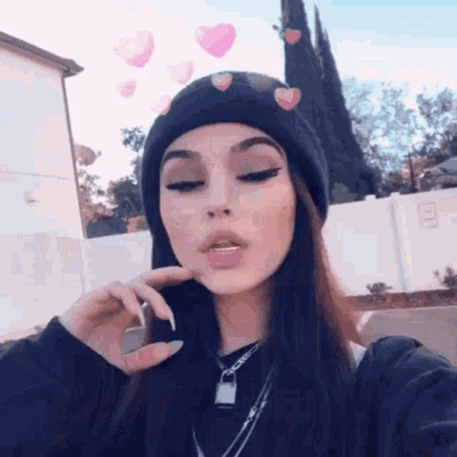 a girl wearing a black beanie and a necklace is taking a selfie with hearts on her face .
