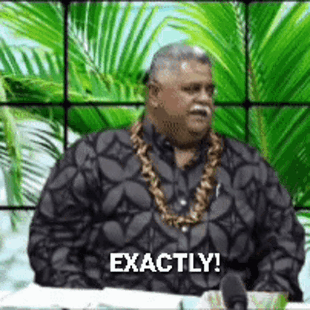 a man wearing a necklace and a black shirt is sitting in front of a screen that says exactly !