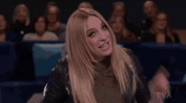 a woman in a leather jacket is sitting in front of a crowd of people and making a funny face .