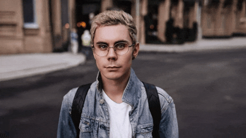 a young man wearing glasses and a denim jacket stands on a street