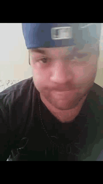 a man with a beard wearing a blue hat and a black shirt is making a funny face .