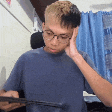 a young man wearing glasses and a blue shirt holds his hand to his head