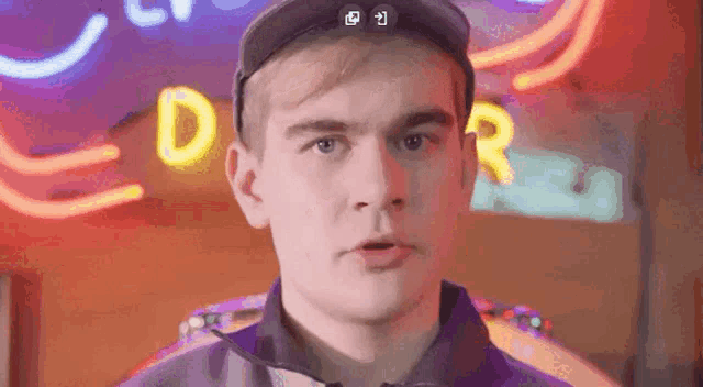 a man wearing a hat and a purple shirt is standing in front of a neon sign with the letter d on it .
