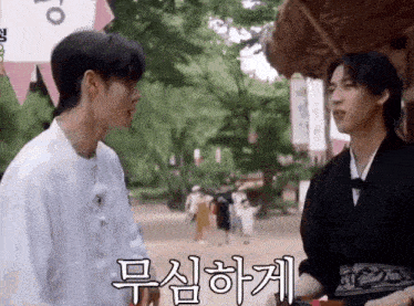 two young men are standing next to each other in a park and talking in korean .