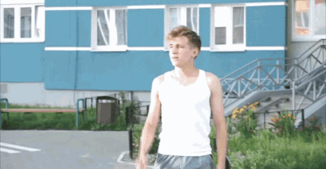 a young man wearing a white tank top stands in front of a blue building