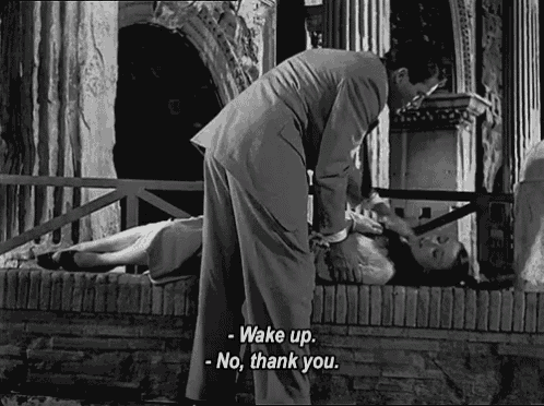 a man in a suit stands next to a woman laying on the ground and says " wake up "