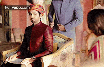 a man in a turban is sitting in a chair while a man in a suit stands behind him .