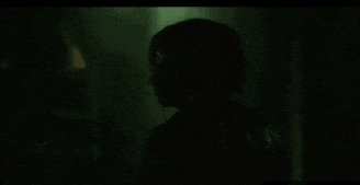 a man in a hooded jacket is standing in a dark room with a green light behind him .
