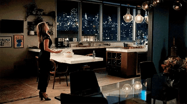 a woman in a black dress is standing in a kitchen with a large window overlooking the city at night .