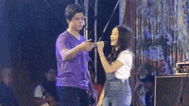 a man in a purple shirt is pointing at a woman in overalls on a stage .