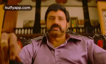a man with a mustache is sitting in a chair with his hand up .