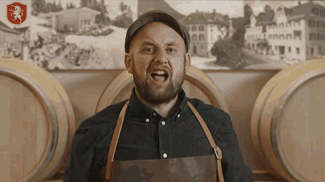 a man with a beard wearing an apron stands in front of barrels