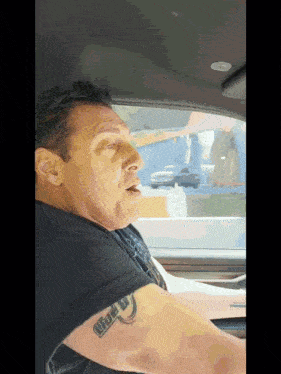 a man in a car has a tattoo on his arm that says " life is "