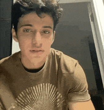 a young man is wearing a brown t-shirt with a fan on it and looking at the camera .