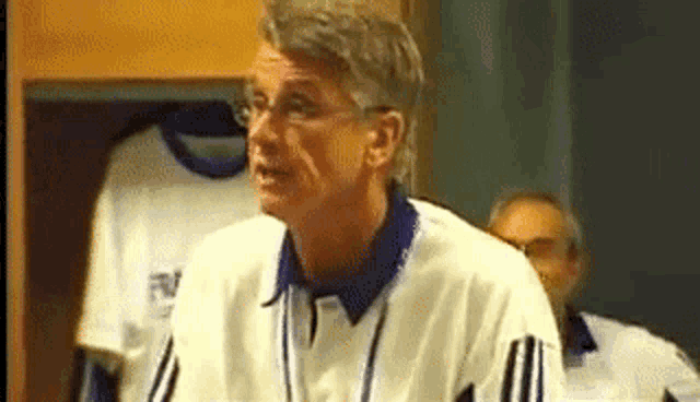 a man in a white shirt with a blue collar is standing in a locker room talking to someone .